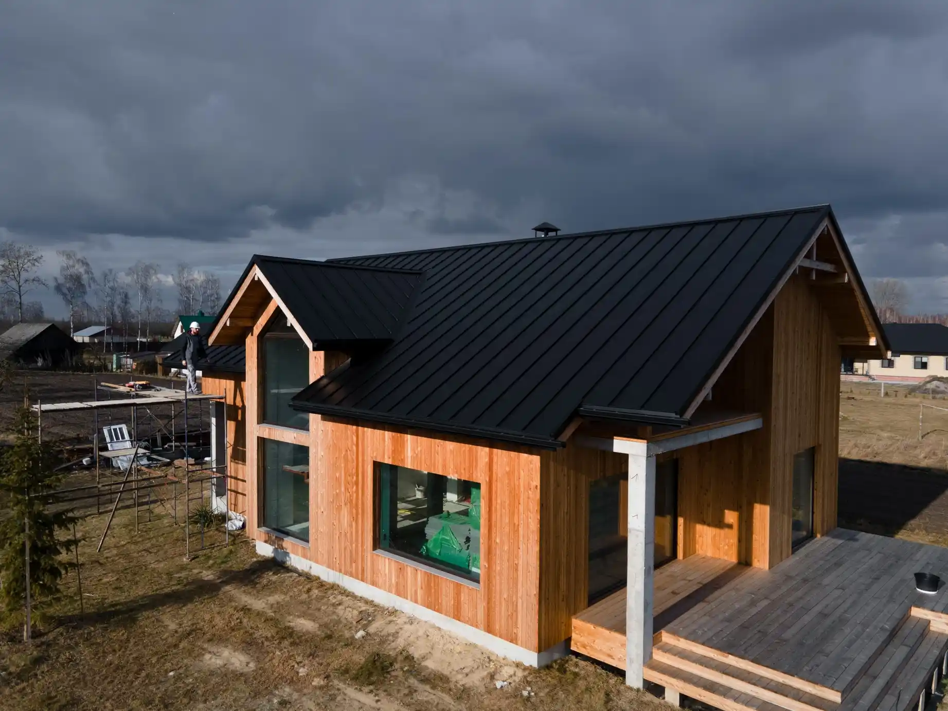Aerial view of a modern wooden house under construction, featuring large windows, a wide wooden deck, and a newly installed black metal roof with a worker standing on scaffolding.