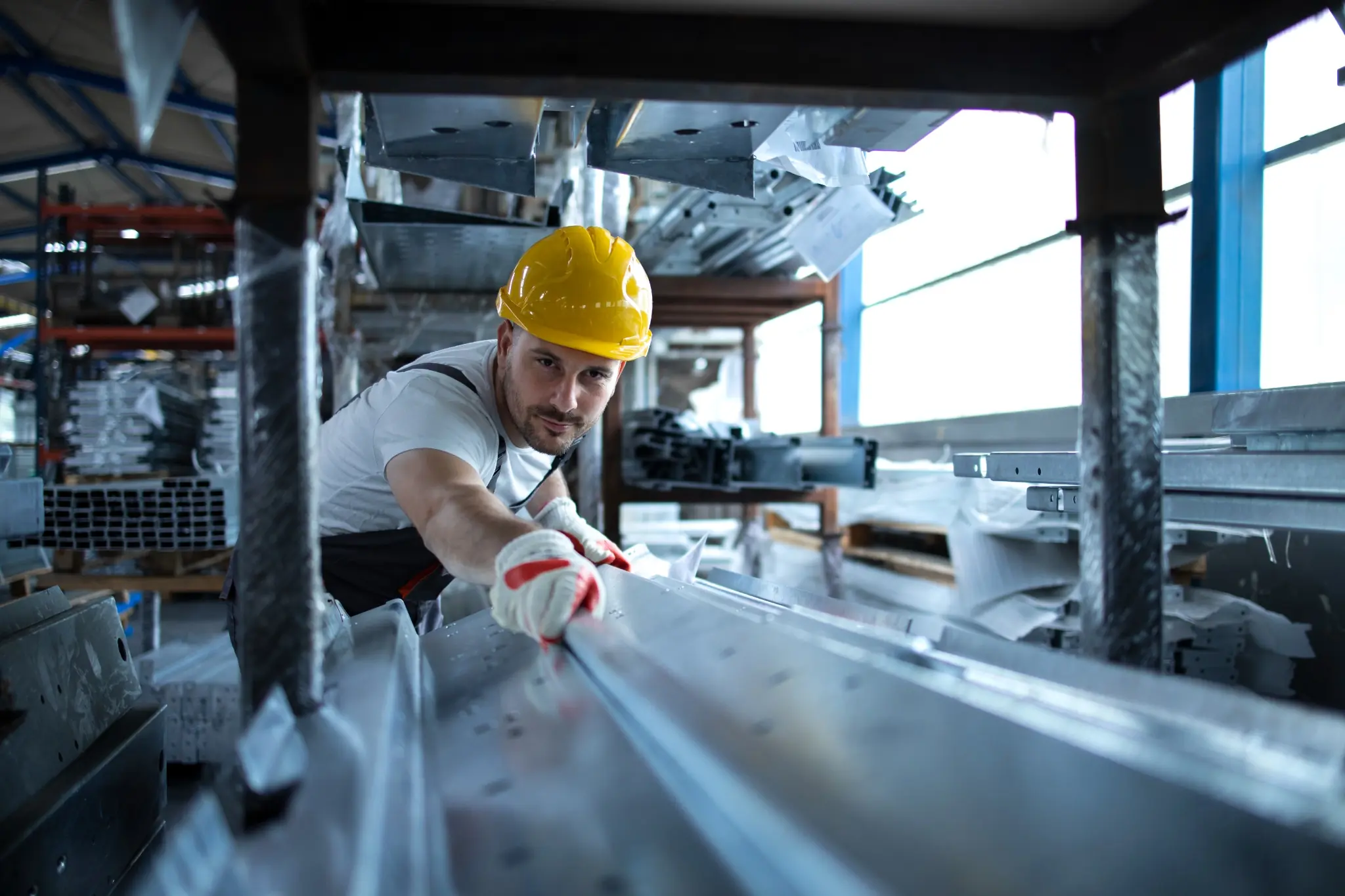A male factory worker wearing a yellow hard hat and safety gloves leans forward to handle large sheet metal components inside an industrial manufacturing warehouse.