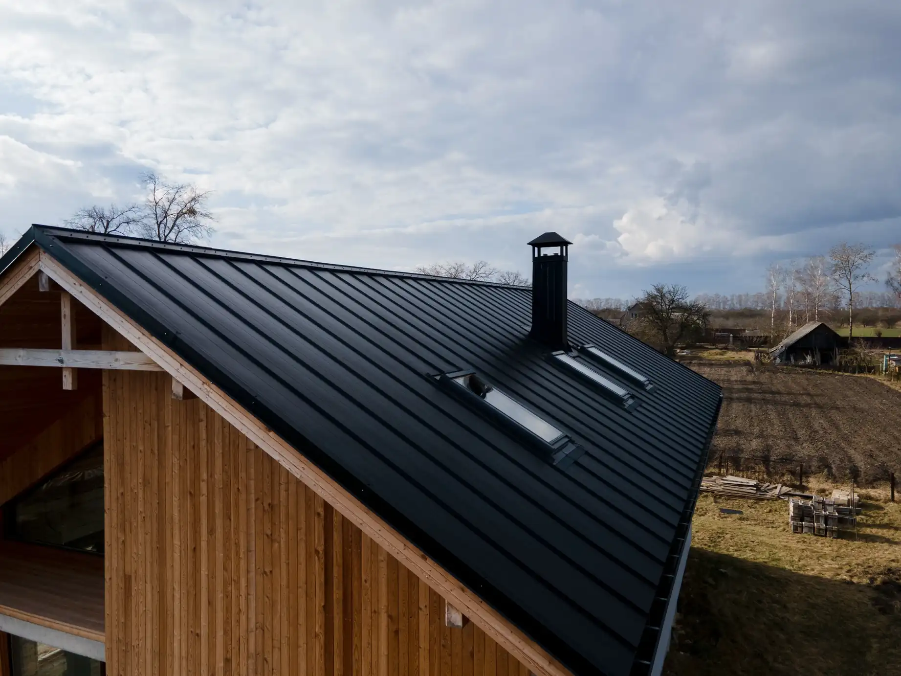 High-angle view of a modern wooden house featuring a sleek black standing seam metal roof with skylights and a chimney, set against a cloudy sky in a rural landscape.