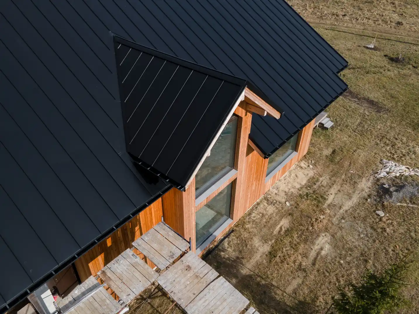 High-angle aerial view of a modern wooden house under construction, showcasing a newly installed black standing seam metal roof, large windows, and exterior wooden scaffolding.