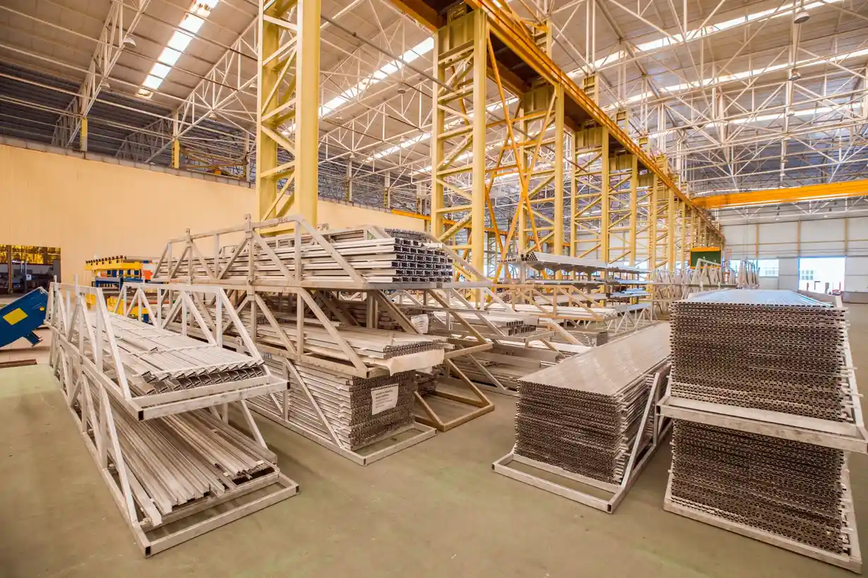 Interior view of a spacious industrial warehouse featuring a yellow steel framework and multiple heavy-duty racks storing stacked metal profiles and construction materials.