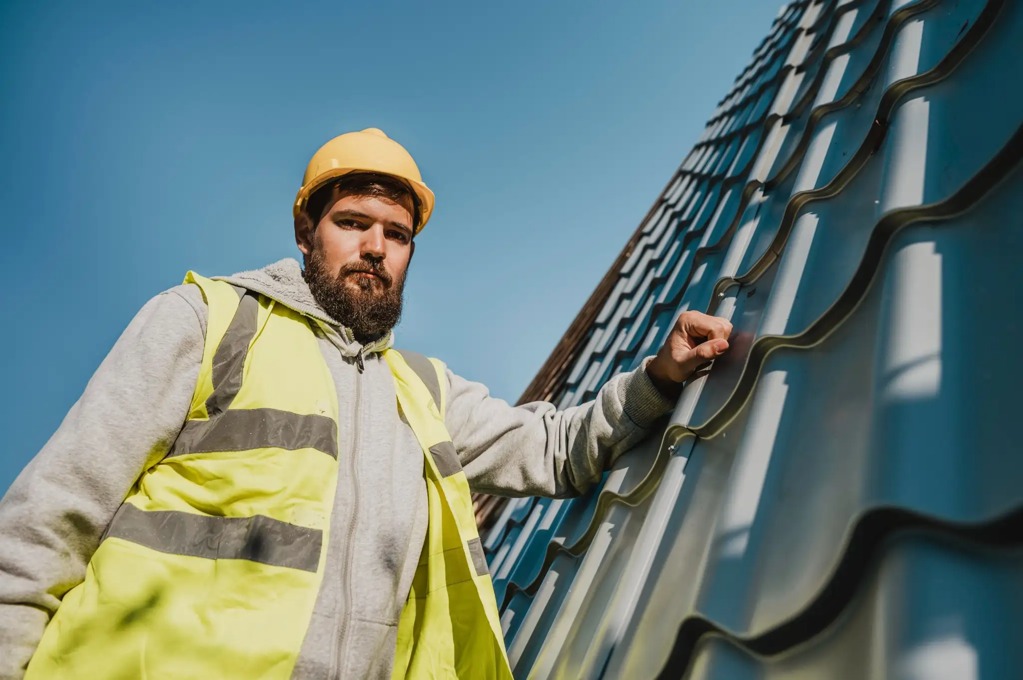 Low-angle portrait of a bearded construction worker wearing a yellow hard hat and high-visibility safety vest standing next to grey corrugated metal roofing panels.