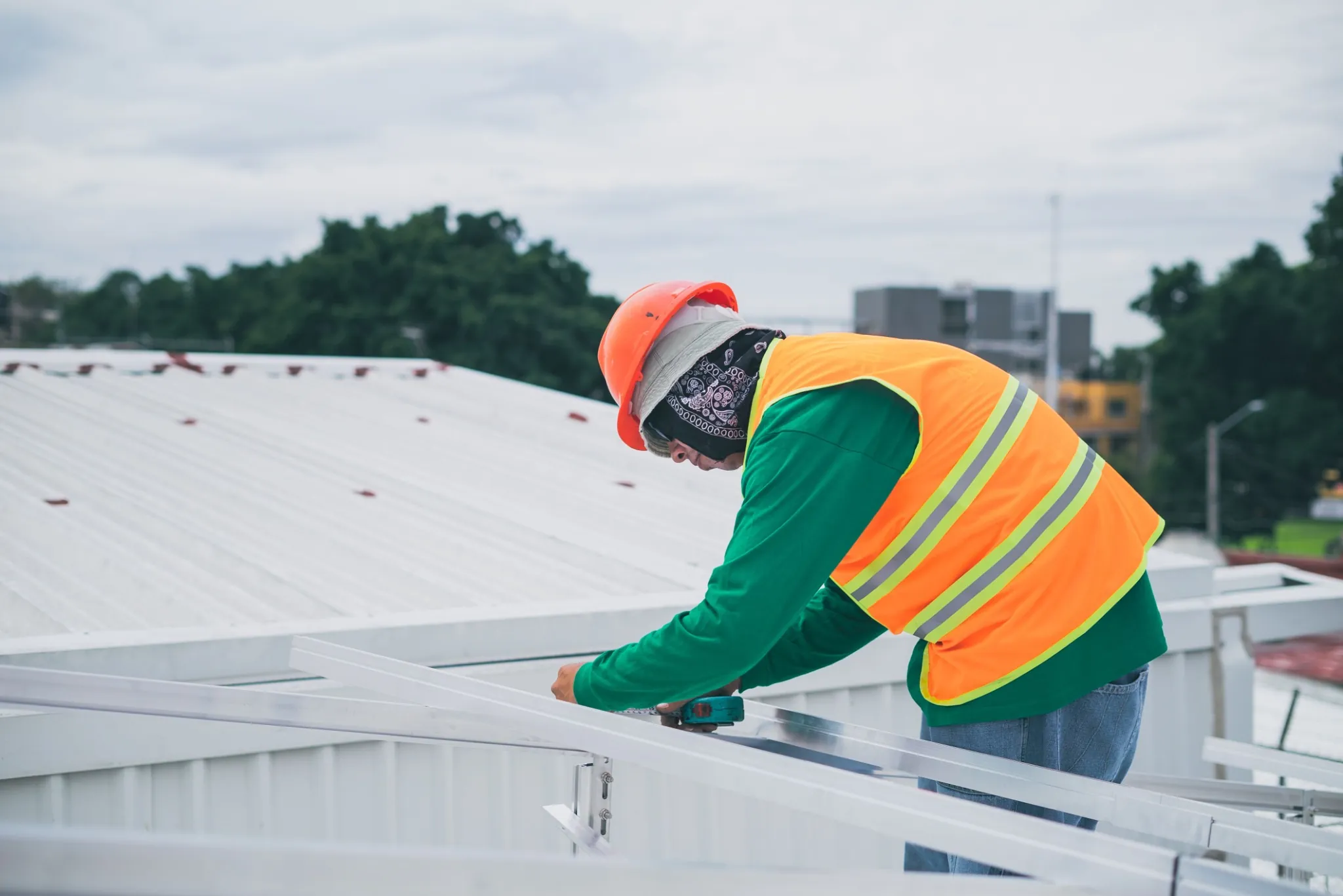 A maintenance worker wearing safety gear, including an orange hard hat and high-visibility vest, assembling metal framing on a commercial white metal roof.
