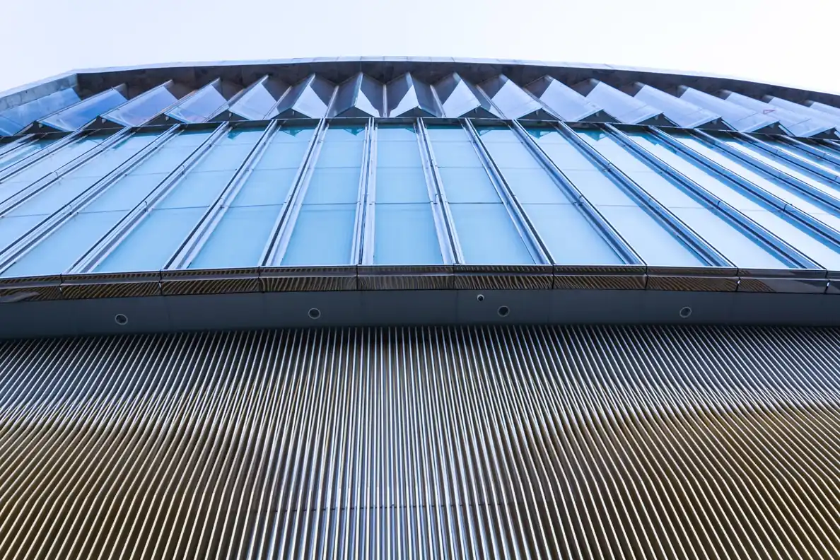 Low-angle architectural view of a modern stadium exterior, showcasing a geometric facade with large blue glass panels above vertical metallic louvers.