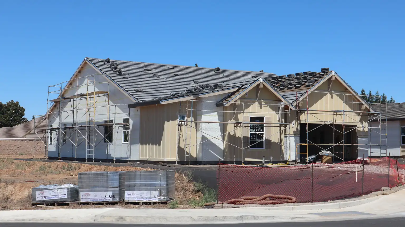 A new residential home under construction in Elk Grove, California, featuring exterior scaffolding, house wrap, and a partially completed roof with stacked roofing tiles ready for installation.