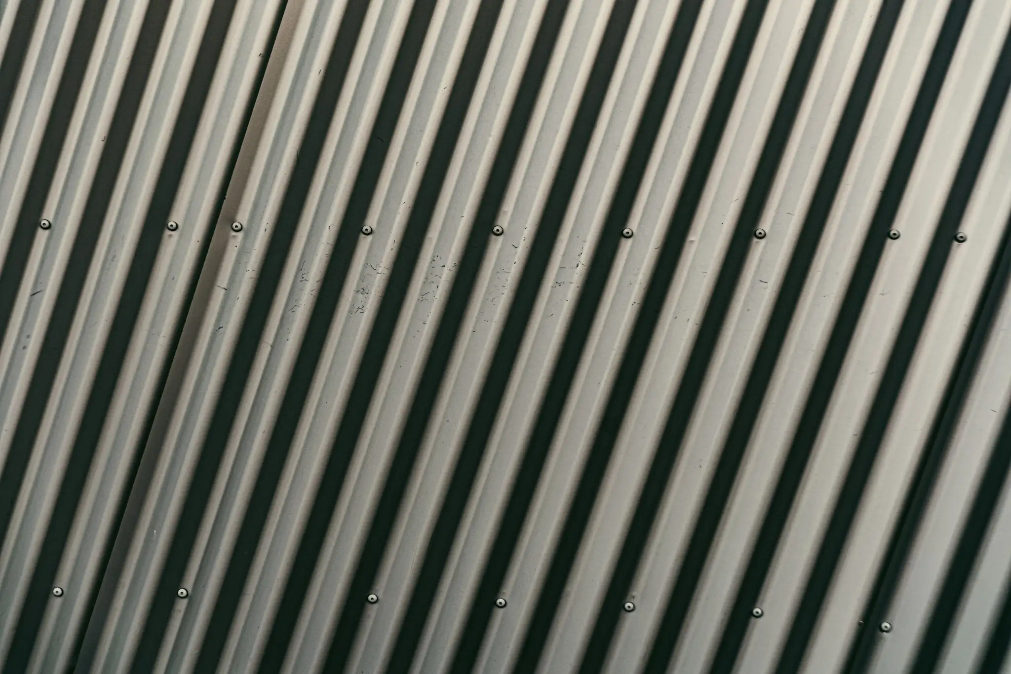 Close-up texture of grey corrugated metal roofing or siding, displaying repeating diagonal ridges and small metal screws.