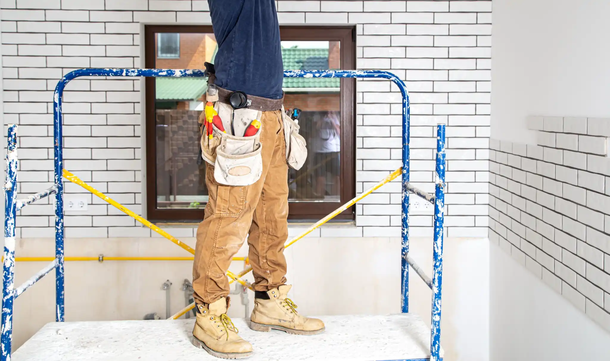 A close-up view of a construction worker wearing a loaded leather tool belt and tan work boots, standing on blue indoor scaffolding during a home renovation project.