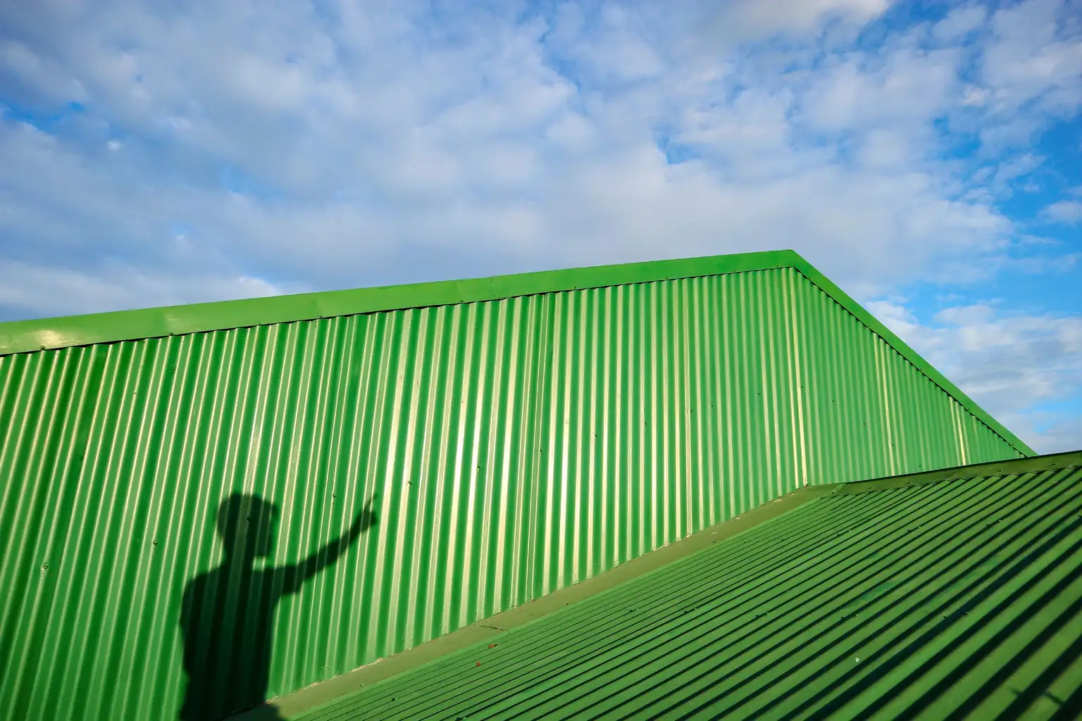The shadow of an anonymous person pointing upward, cast against a bright green corrugated metal roof and building exterior under a sunny blue sky.