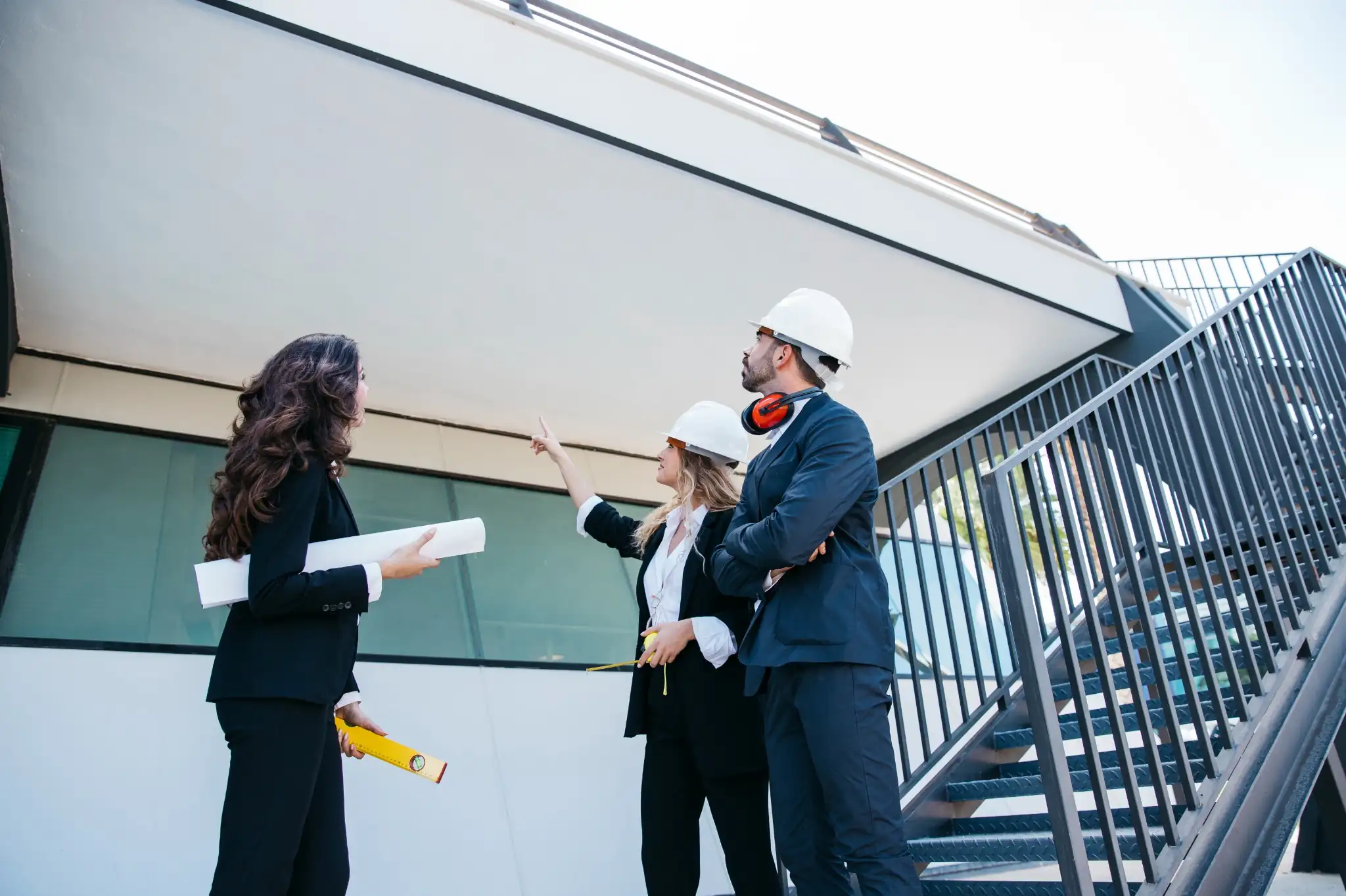A team of three architects and project managers in business attire and white hard hats inspecting a commercial building site, discussing the structure near an outdoor metal staircase with blueprints and a level in hand.