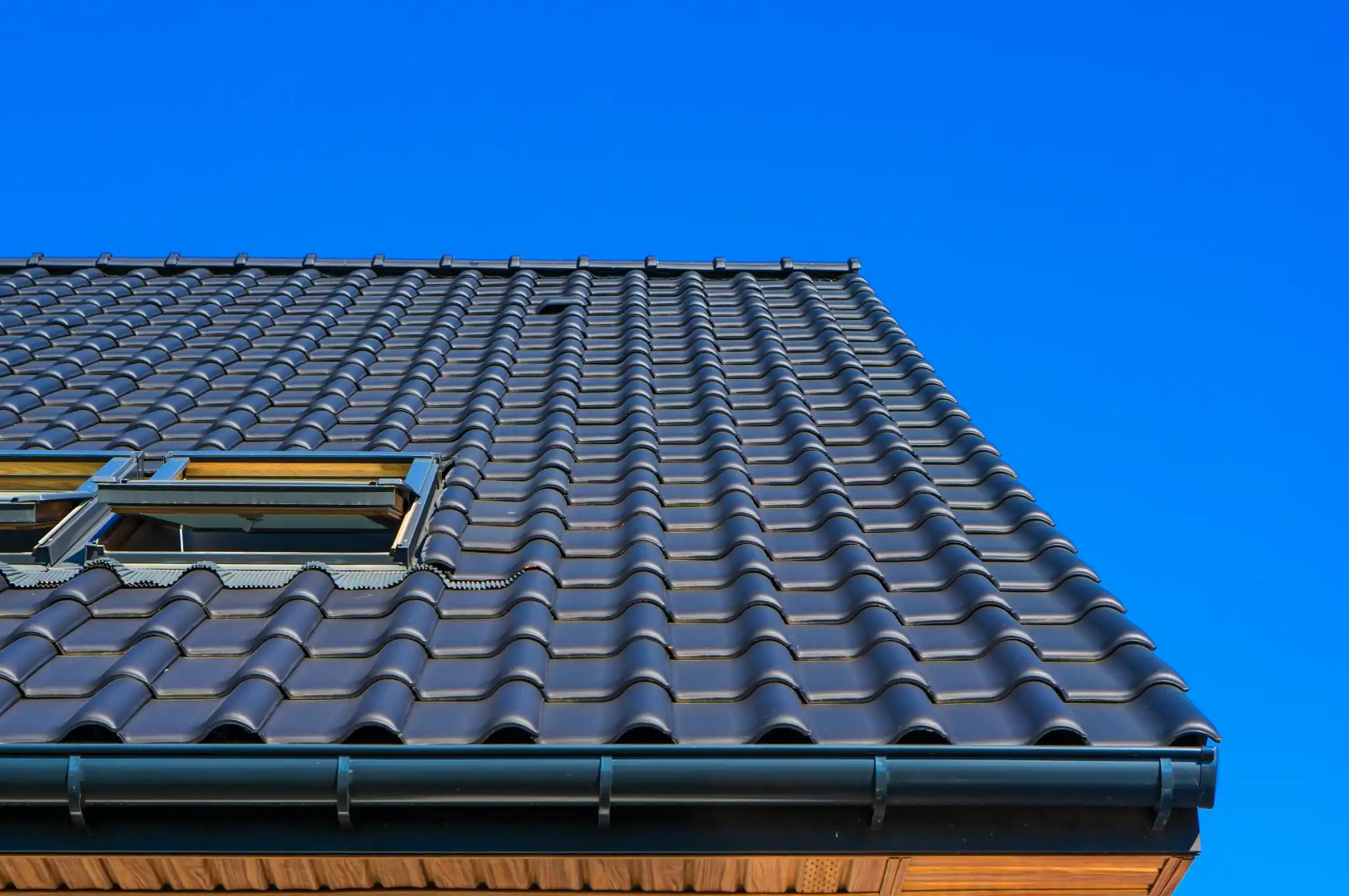 A low-angle close-up shot of a modern residential black tile roof featuring open skylights and dark gutters, set against a clear, vibrant blue sky.