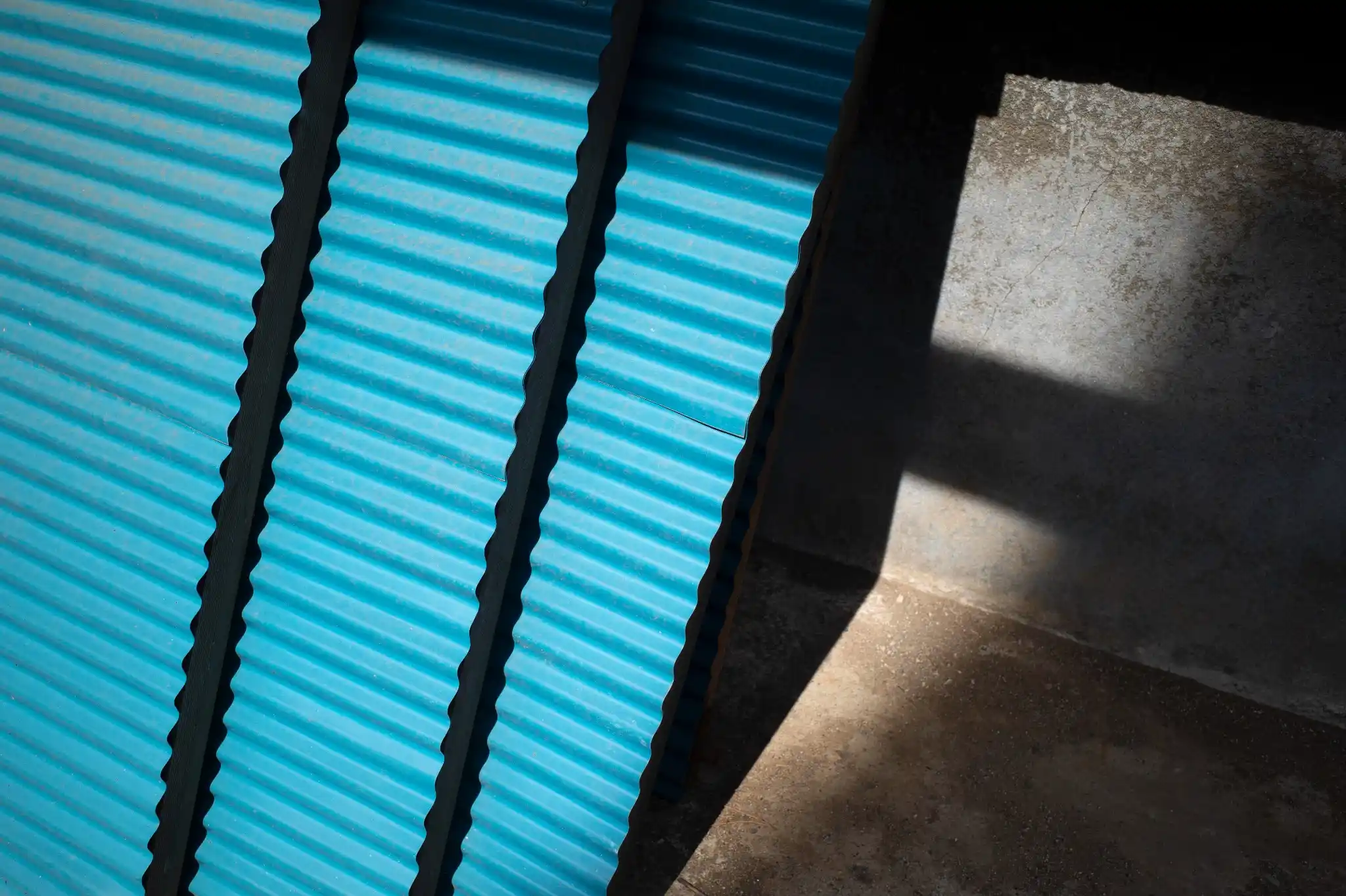 An abstract outdoor view of bright blue corrugated metal roofing sheets leaning against a textured concrete wall, casting sharp diagonal shadows in the sunlight.