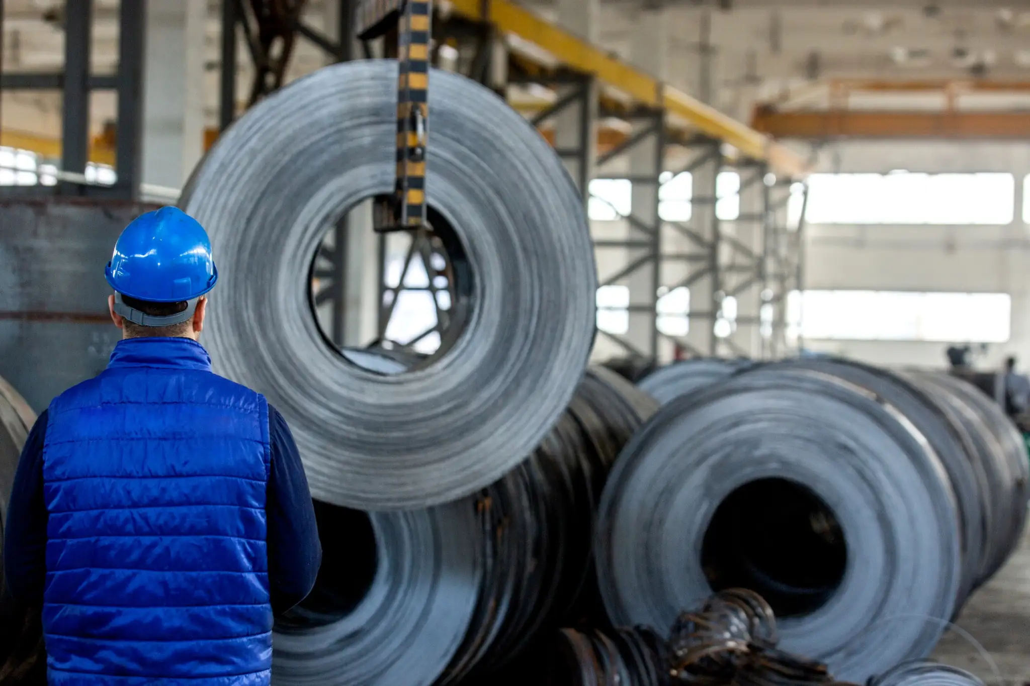 An industrial worker wearing a blue hard hat and safety vest stands in a manufacturing plant, observing a massive steel metal coil being lifted by an overhead crane.